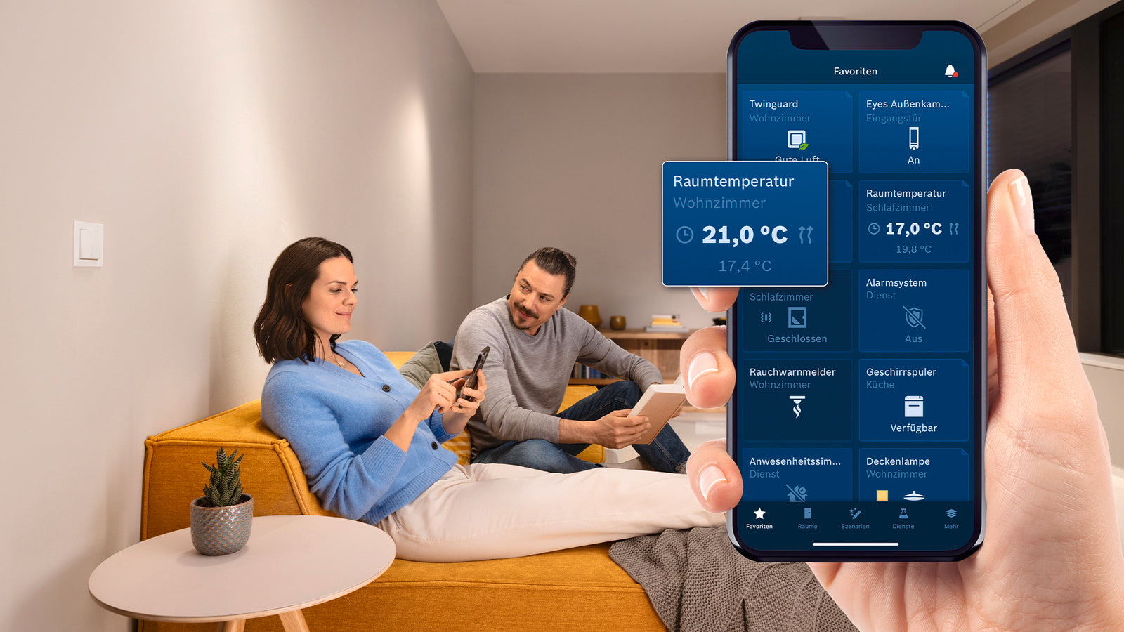 Bosch Smart Home App auf dem Smartphone mit Kachel Raumtemperatur 21,0 Grad, Paar auf dem Sofa dahinter