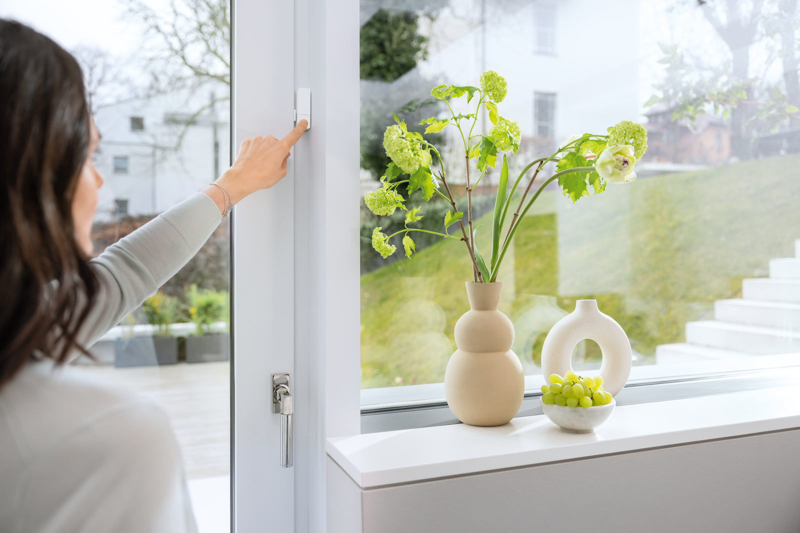 Frau drückt den Button des Tür-/Fensterkontakt II [+M] am Fensterrahmen, Fensterbank mit Vasen daneben