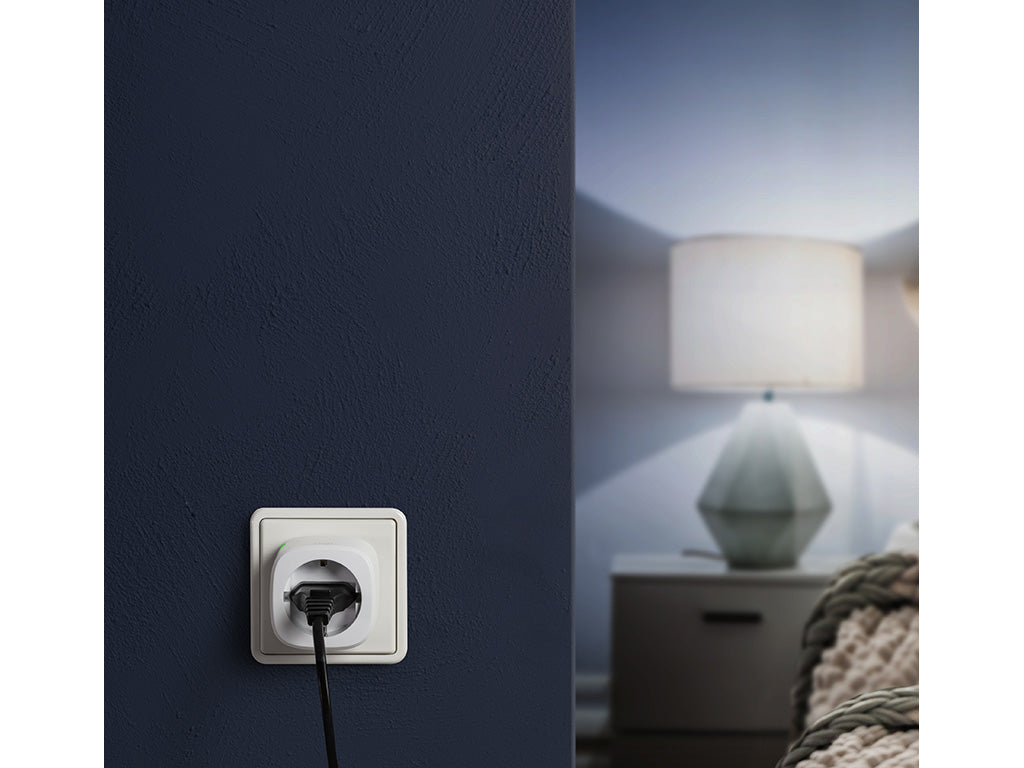 Eve Energy in einer Steckdose an dunkelblauer Wand, dahinter leuchtet eine Nachttischlampe