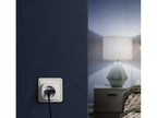 Eve Energy in einer Steckdose an dunkelblauer Wand, dahinter leuchtet eine Nachttischlampe