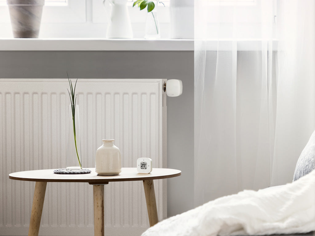 Eve Thermo Control auf einem Tisch im Schlafzimmer, dahinter Heizkörper mit Eve Thermo am Ventil