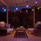 Nanoleaf Outdoor String Light bunt leuchtend unter einer Holzpergola über zwei Sesseln und Couchtisch bei Nacht