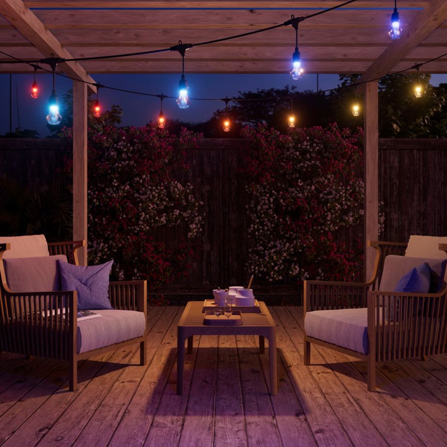 Nanoleaf Outdoor String Light bunt leuchtend unter einer Holzpergola über zwei Sesseln und Couchtisch bei Nacht