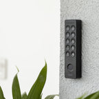 Nuki Keypad 2 an einer weiß verputzten Außenwand neben der Tür montiert, Pflanzenblätter im Vordergrund