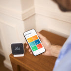 Mann hält Smartphone mit der tado App, dahinter der Funk-Temperatursensor X auf der Kommode