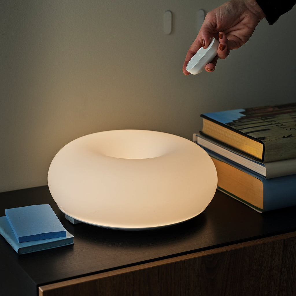 IKEA VARMBLIXT leuchtet warmweiß auf einem dunklen Sideboard, eine Hand richtet die Fernbedienung auf die Leuchte