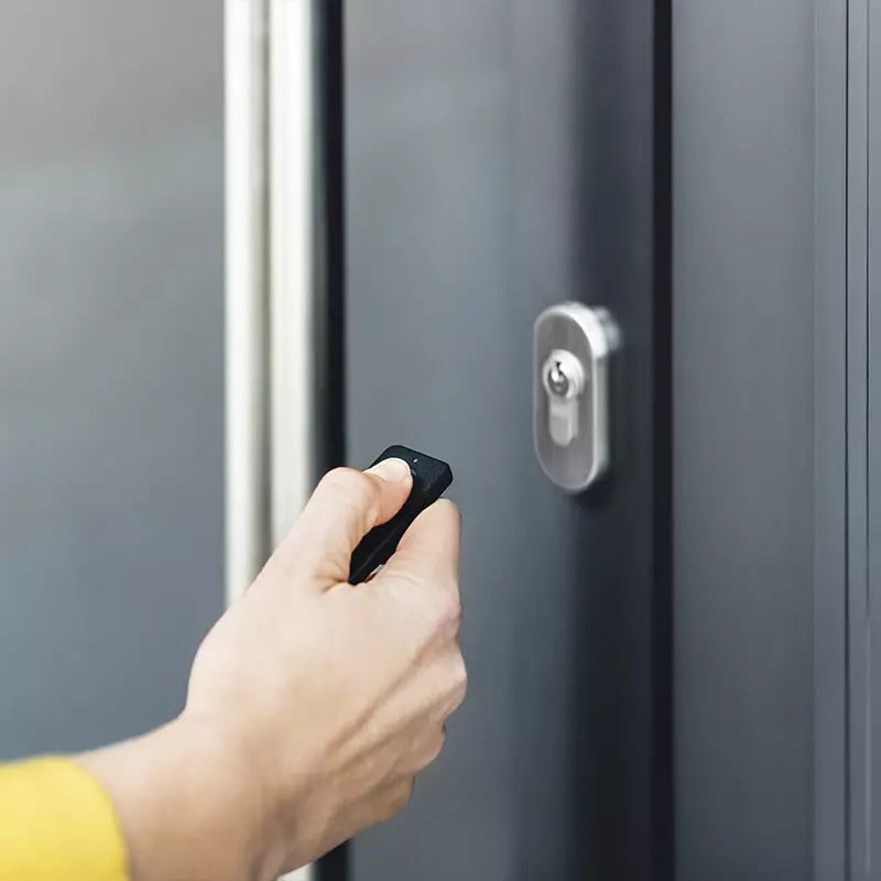 Hand hält den Nuki Fob mit dem Daumen auf der Taste vor eine dunkelgraue Haustür