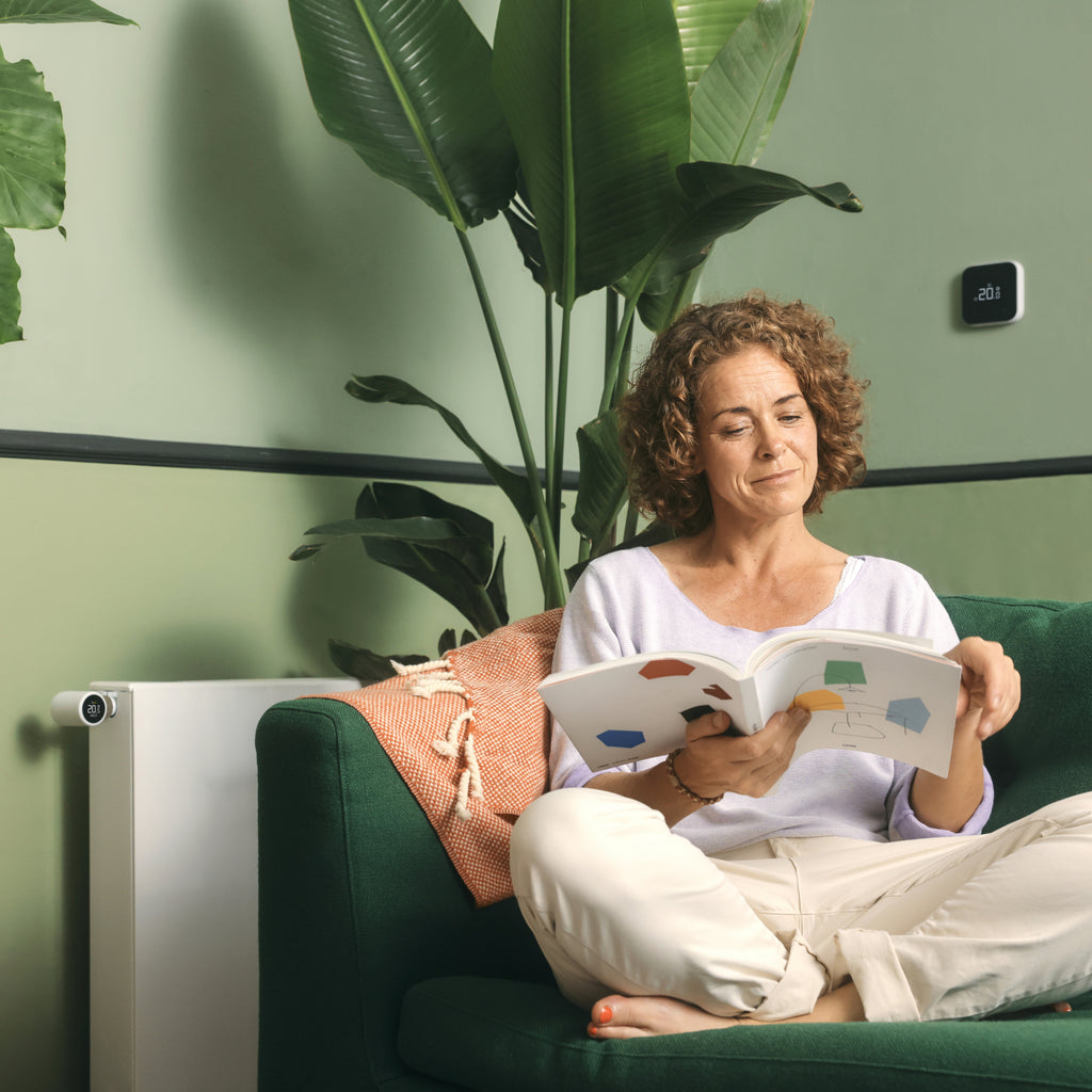 Frau liest auf dem Sofa, an der Wand der Funk-Temperatursensor X, links am Heizkörper ein Thermostat X