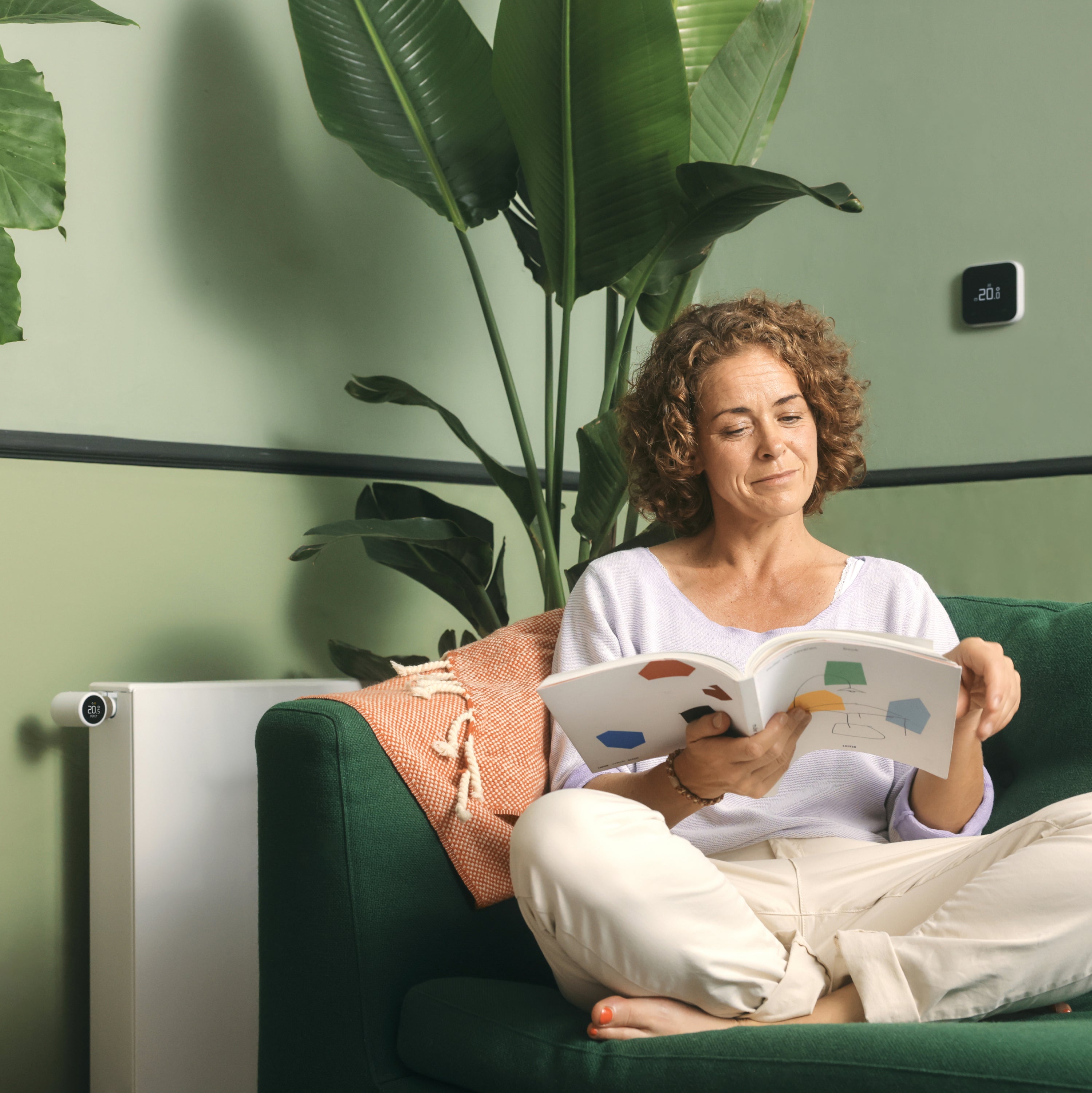 Frau liest auf dem Sofa, an der Wand der Funk-Temperatursensor X, links am Heizkörper ein Thermostat X