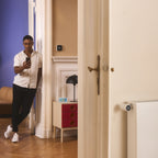 Mann mit Smartphone im Türrahmen, Thermostat X an der Wand im Nebenraum, Heizkörperthermostat am Radiator