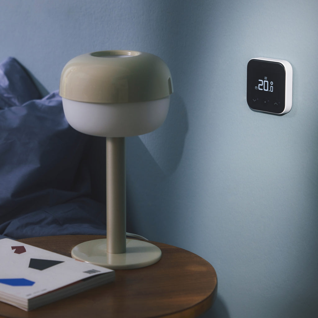 Thermostat X leuchtet an der blauen Schlafzimmerwand, davor Nachttischlampe und aufgeschlagenes Buch